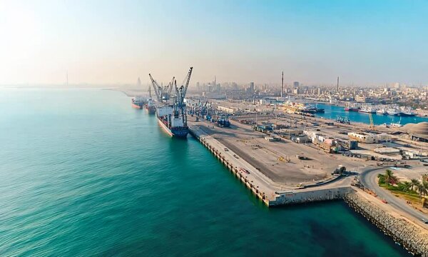 Aerial view of a seaport and industrial area in the port of Al Khalifa, Saudi Arabia. The harbor landscape with ships docked along the seacoast, with a mountainous background.