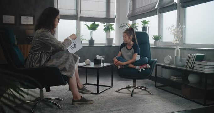Female child psychologist shows illustrations to Asian school-aged girl, provides psychological test during therapy. Girl sits in chair and looks at pictures. Kid's counseling session. Psychoanalysis.