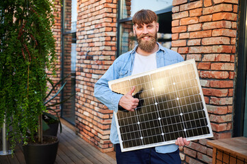 Portrait of bearded man holds solar panel, giving thumbs up, standing in front of brick building...