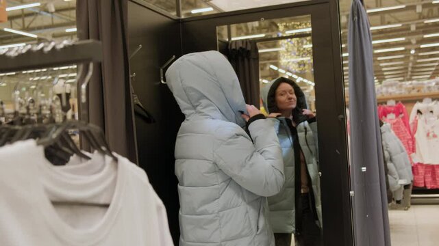 Woman trying on jacket in front of mirror in fitting room before making purchase in clothing store in shopping mall.