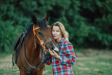 Obraz premium Happy blonde with horse in forest. Woman and a horse walking through the field during the day. Dressed in a plaid shirt and black leggings.