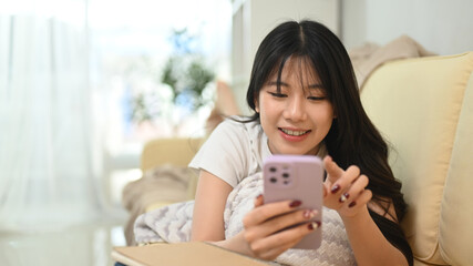 Smiling young woman texting message and scrolling news in social media app on her mobile phone