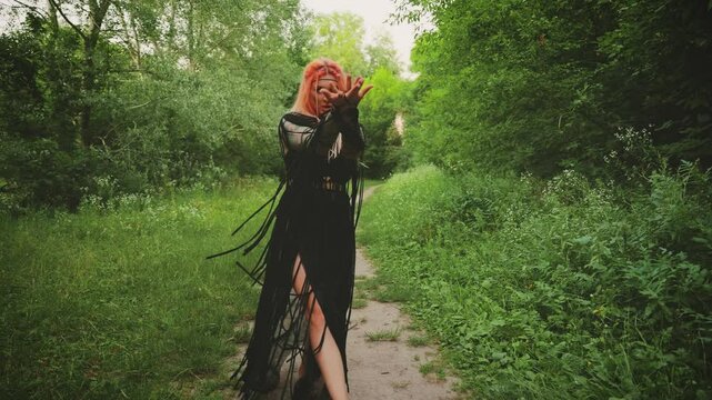 Halloween holiday woman witch conjuring with hands, dancing looking at camera walking in dark forest green tree. long black dress gothic boho style macrame, elf fairy girl shaman. orange colored hair.