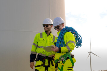 Two engineers in high visibility gear stand near a wind turbine, reviewing a clipboard and preparing for maintenance work. The image highlights teamwork, safety, and renewable energy sector.