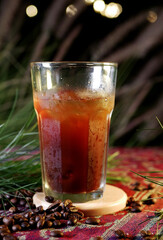 Iced Americano in a large glass with visible refreshing ice cubes, placed on a wooden coaster. Coffee beans are scattered on colorful fabric, and the background shows bokeh lights and green leaves.