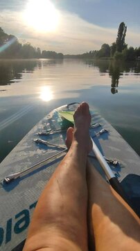 Bare legs of woman on SUP board on surface of river or lake with sun reflecting and with trees on banks on warm summer evening. Person swimming on SUP board on pond on calm day. Tourism rest sport