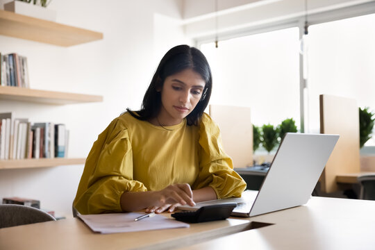 Focused young Indian businesswoman calculating budget, counting income, planning expenses. Accountant, entrepreneur analyzing financial data, investment using calculator at laptop in office