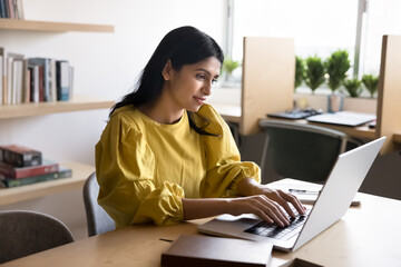 Positive young Indian professional girl typing on laptop in co-working room working at computer,...