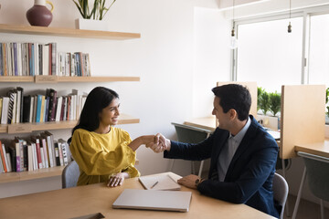 Fototapeta premium Confident Hispanic businessman hiring young Indian Intern woman for job, shaking hand with new employee after interview meeting, giving handshake over large table, getting agreement