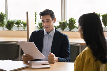 Positive young Hispanic businessman interviewing job candidate woman, reading paper resume at...