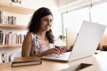 Happy young Indian project manager woman typing on laptop at workplace table, working at computer, looking at screen, smiling, enjoying online job business communication