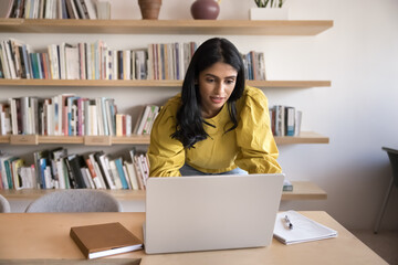 Focused busy young Indian professional woman standing at workplace table, leaning over laptop,...