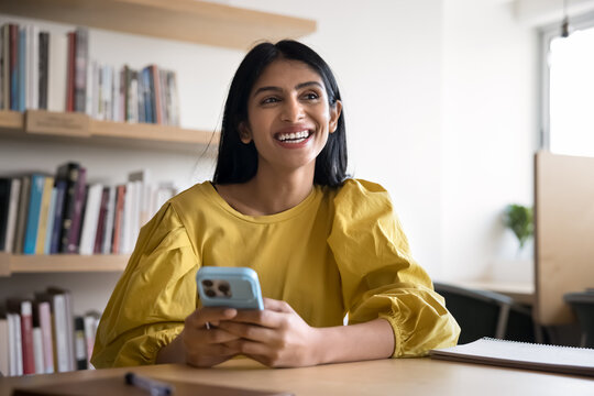 Happy dreamy young Indian student woman holding mobile phone and looking away, sitting at table in library, smiling, laughing, using smartphone for Internet communication, education