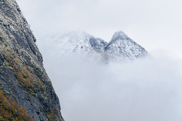 A rocky cliff edge in the foreground with a snowy mountain range in the background. White clouds cover the upper portion of the mountains.
