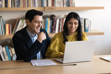 Happy diverse couple of business colleagues working at computer together, discussing online project, marketing strategy, teamwork, watching presentation on laptop in office library