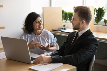 Obraz premium Positive couple of Indian and Caucasian office colleagues discussing job, work tasks, meeting at computer for teamwork, brainstorming, sitting at office table, talking
