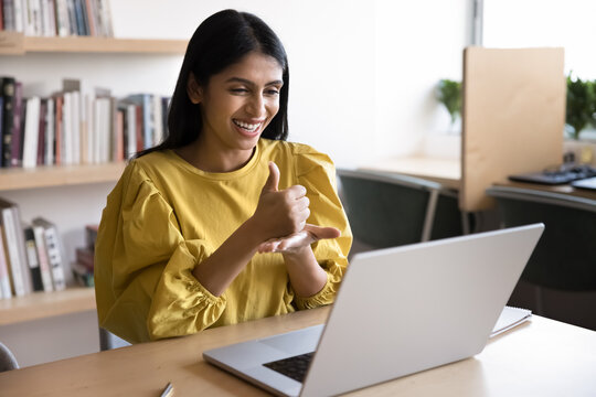 Cheerful young Indian student with hearing disability talking on video call at laptop in library, showing like, thumb up gesture on palm at computer, using sign language for Internet communication