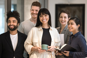 Happy diverse office team with mature female Latin leader standing together, looking away, smiling, laughing, holding notebook, cup. Multiethnic business colleagues posing shooting