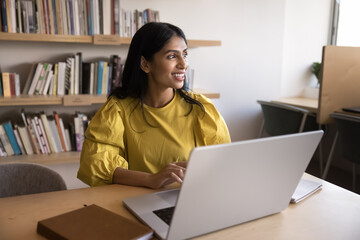 Happy thoughtful young Indian professional woman working on research study in library, using laptop at table, looking away, smiling, thinking on successful business project solution