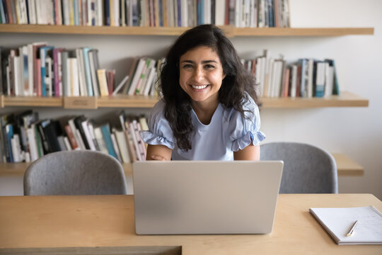 Happy beautiful Indian student woman working at laptop in library, posing for portrait at workplace table, looking at camera, bending over desktop, smiling, laughing