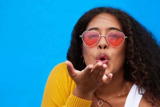 Woman, portrait and blow kiss in studio with heart sunglasses for valentine day, flirt gesture and romance. Girl, eyewear and love emoji, date inspiration and gratitude connection on blue background - Powered by Adobe
