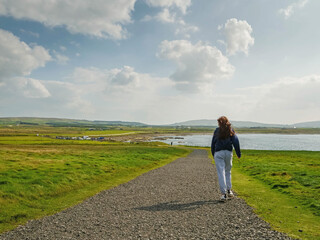Teenager girl walking on a small country road. Stunning nature landscape in the background with green fields, blue ocean, mountains and cloudy sky. Downpatrick head, Ireland. Travel and tourism.