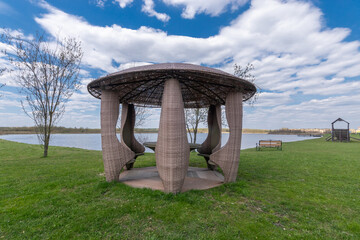 Gazebo with a round roof and original shaped seats near a small pond, against a background of...