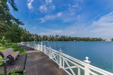Beautiful summer landscape with a picturesque lake against the blue sky. The shore with lush greenery is decorated with a wooden embankment.