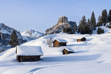 Evening light illuminates wooden cabins in a snow covered valley with the famous Sassongher mountain in the background. The peaceful winter landscape feels almost magical