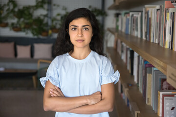 Serious beautiful young Indian bookstore manager posing with arms crossed at bookshelves, looking...