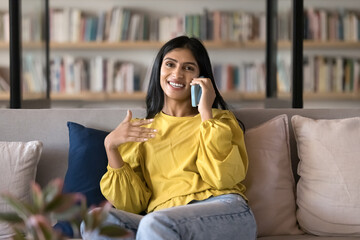 Positive beautiful young Indian woman talking on mobile phone on comfortable couch. Student making...