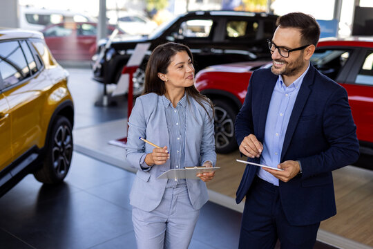 Super sales team in dealership, two consultants or managers in elegant suits with laptop and tablet in arms in car dealership