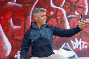 Gray haired man in his 40's smiles as he takes a selfie against a green graffiti wall, wearing a blue raincoat. Urban and casual street style atmosphere