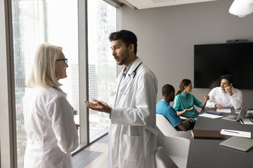 Fototapeta premium Young male doctor multiethnic clinic staff member speak to older female colleague at hospital boardroom. Medic intern talk with mid aged mentor experienced physician ask advice consult about diagnosis