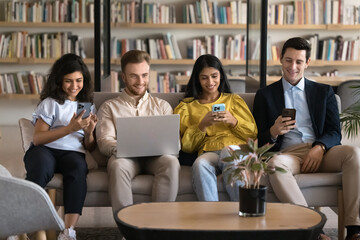 Cheerful multiethnic team of four student friends focused on gadgets sitting on couch in library together, using digital devices, browsing internet, smiling, laughing, ignoring each other