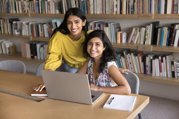 Happy Indian young adult female student friends studying in library together, meeting at laptop computer, cooperating on learning project together, looking at camera, smiling, posing for portrait