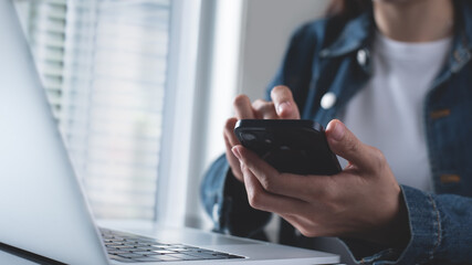 Closeup, business woman sitting at table using mobile smrt phone during working on laptop computer at home office. Young asian woman freelancer  surfing the internet, online working from home