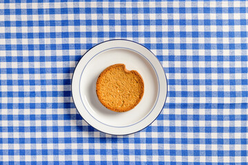 Top view of white ceramic breakfast plate with a typical dutch rusk and a  blue checkered tablecloth as background.