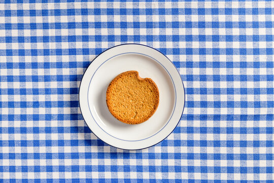 Top view of white ceramic breakfast plate with a typical dutch rusk and a  blue checkered tablecloth as background.