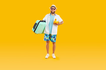 Full body photo of a funny young happy man tourist with suitcase and inflatable duck going on summer holiday trip in beach clothes isolated on studio yellow background. Vacation and travel concept.