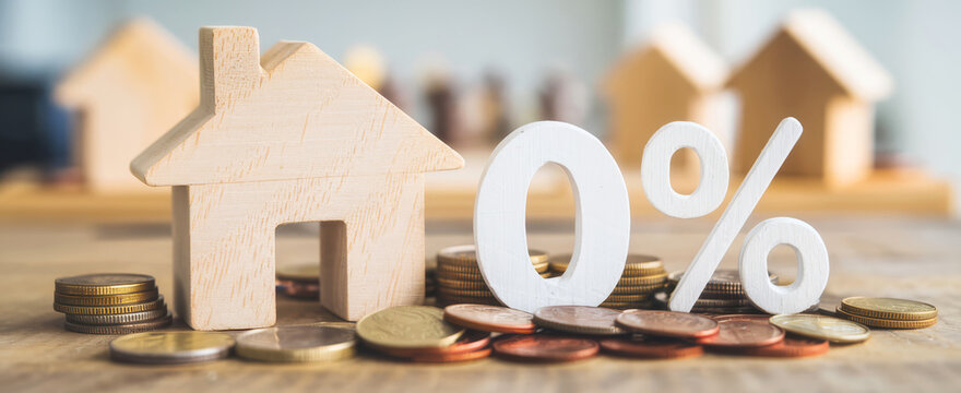 A wooden house model stands beside coins and a "0 percent" sign, symbolizing low mortgage rates or affordable housing options.