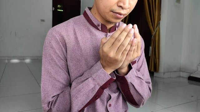 Muslim man wearing red koko shirt raises his hands and prays until finished inside home