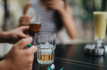 A close-up of a refreshing drink with ice cubes in a glass on a table, surrounded by people enjoying beverages in a relaxed cafe atmosphere.