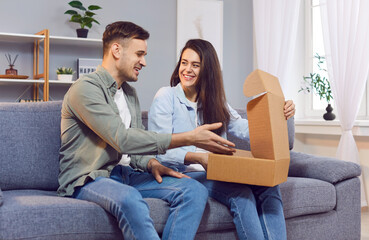 Happy young married couple unpacking parcel from online store or marketplace. Smiling man and woman sitting on couch open cardboard box and unwrap what's inside. Online shopping, delivery concept