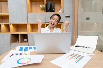 A woman is sitting at a desk with a laptop open in front of her. She is smiling and she is enjoying...
