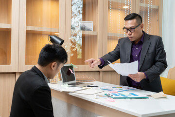 A man in a suit is pointing at a piece of paper on a desk. The man is wearing glasses and he is giving instructions or directions to the other person
