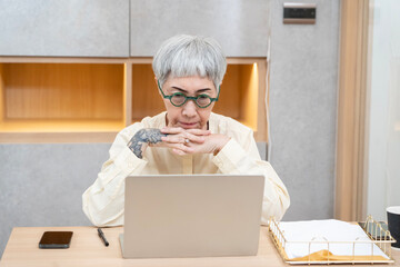 A woman wearing glasses is sitting at a desk with a laptop and a cell phone. She is looking at the...