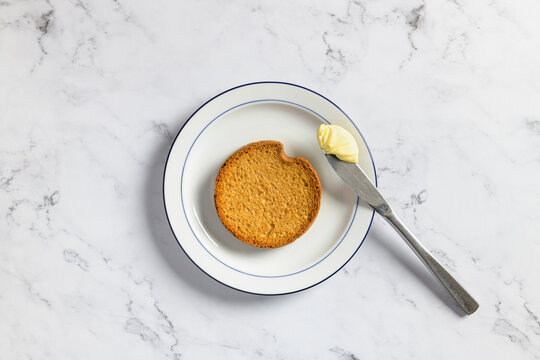 Top view of a traditional dutch rusk and a kitchen knife with creamy butter on a white ceramic plate against a marble background.
