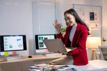 A woman in a red jacket is sitting at a desk with a laptop in front of her