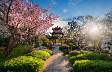 Fototapeta premium A red and green pagoda in a lush garden with cherry blossoms, a stone path, and bright blue sky.